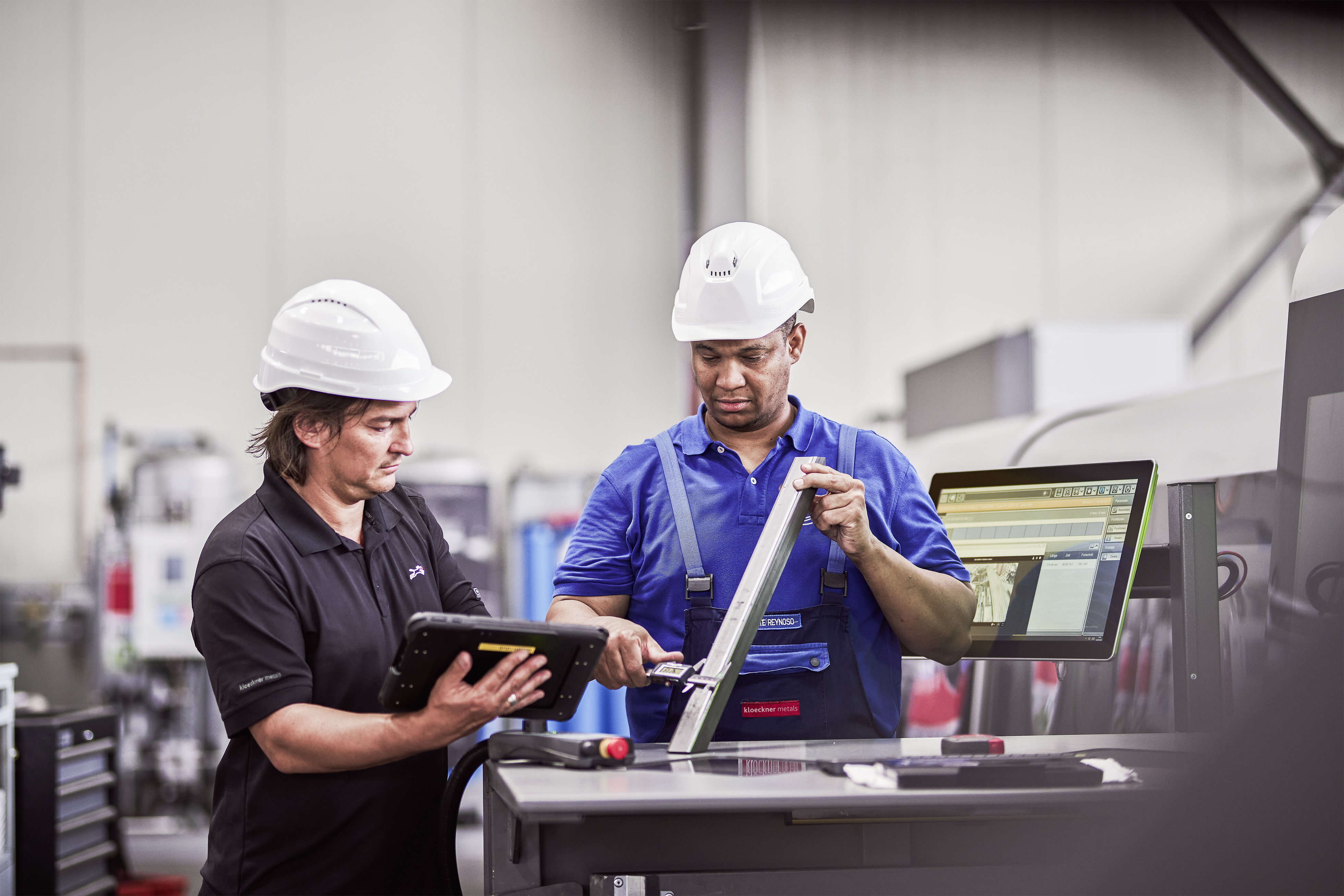 Zwei Arbeiter in einer Produktionshalle tragen Schutzhelme und Arbeitskleidung. Einer hält ein Tablet in den Händen, während der andere mit einem Messgerät ein Metallprofil prüft. Im Hintergrund steht ein Computerbildschirm mit technischen Daten. Die Szene zeigt eine Zusammenarbeit zweier Mitarbeiter bei Kloeckner.