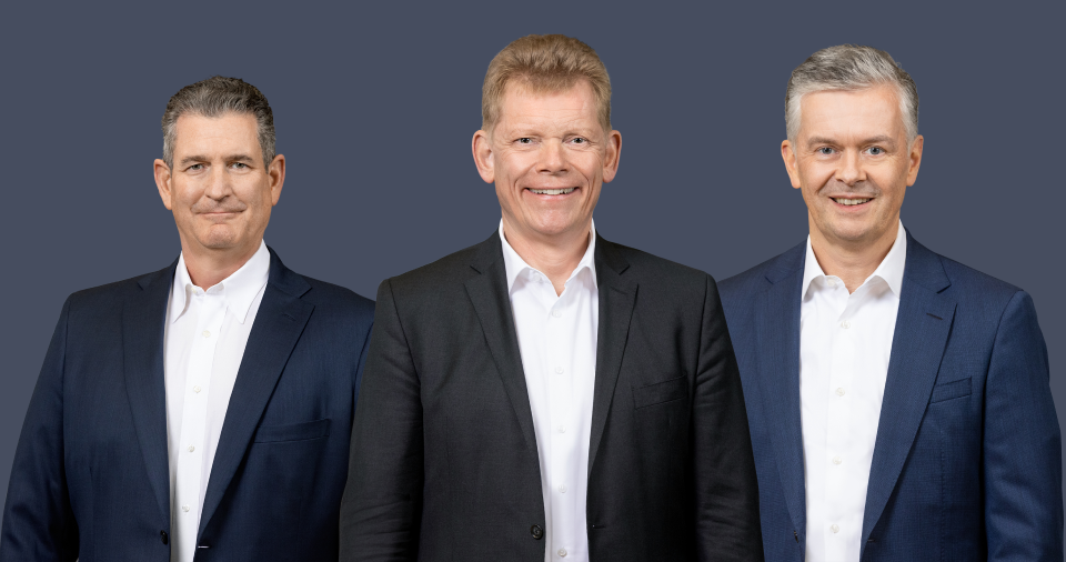 Vor einem dunkelblauen Hintergrund stehen drei Männer in Anzügen und weißen Hemden, die lächeln und nach vorne blicken. Von links nach rechts sind dies John Ganem, Guido Kerkhoff und Dr. Oliver Falk.