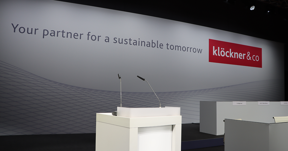 Leere Konferenzbühne mit Podium, Mikrofonen und einer Kulisse mit der Aufschrift „Ihr Partner für eine nachhaltige Zukunft“ und dem Logo von „Klöckner & Co“.