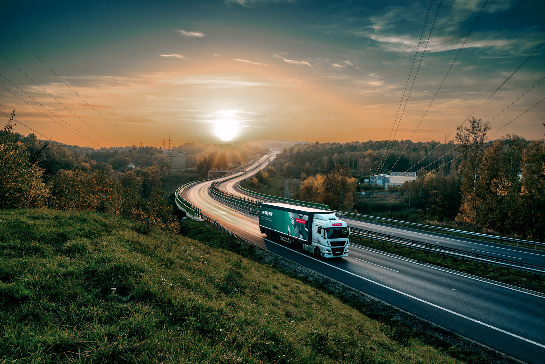 A truck travels on a highway at sunset, with a vibrant sky and blurred headlights creating light trails. Lush greenery lines the road.