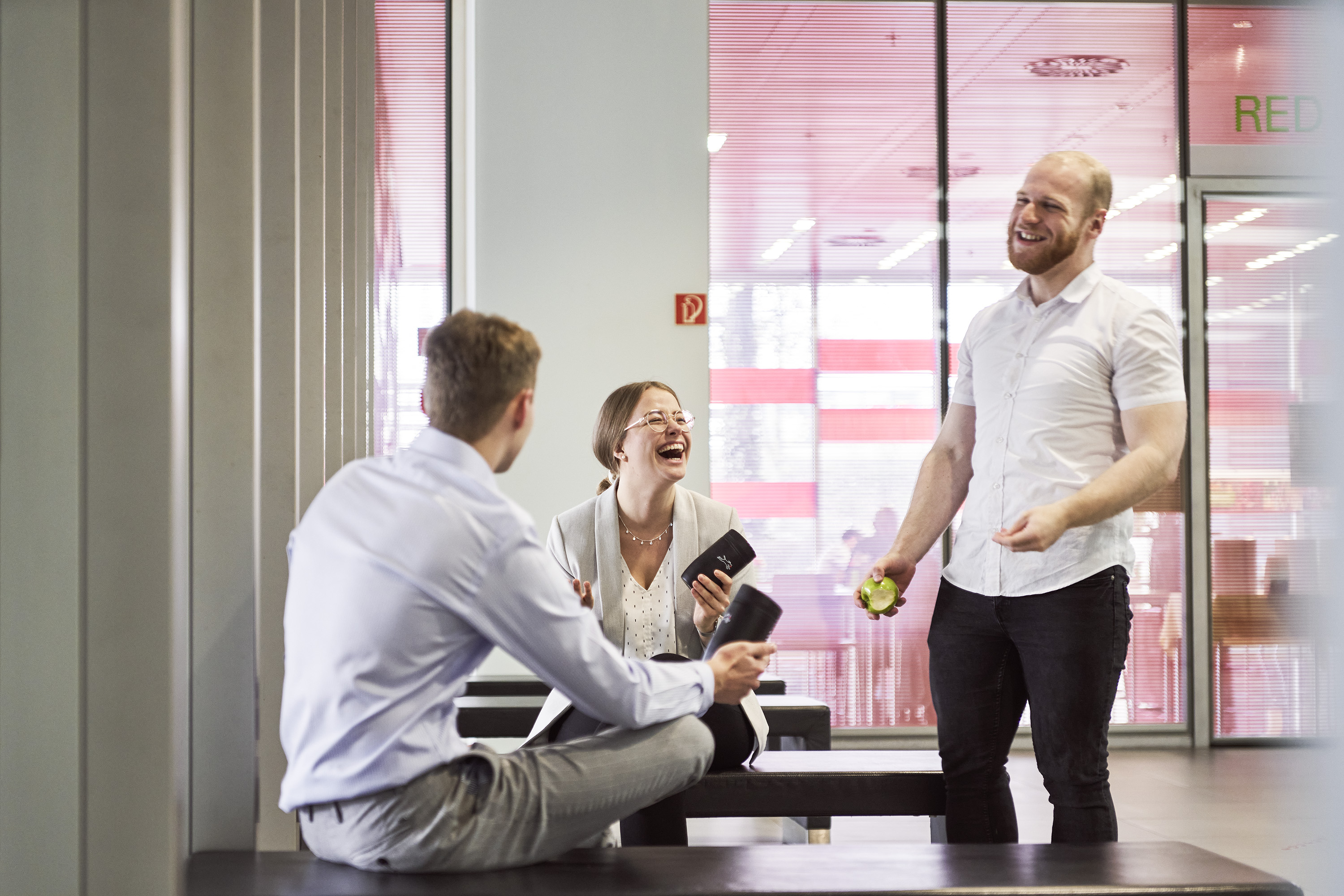 Three people in a modern office lounge, laughing and talking. One holds a phone, another an apple. Glass walls with red accents in the background.