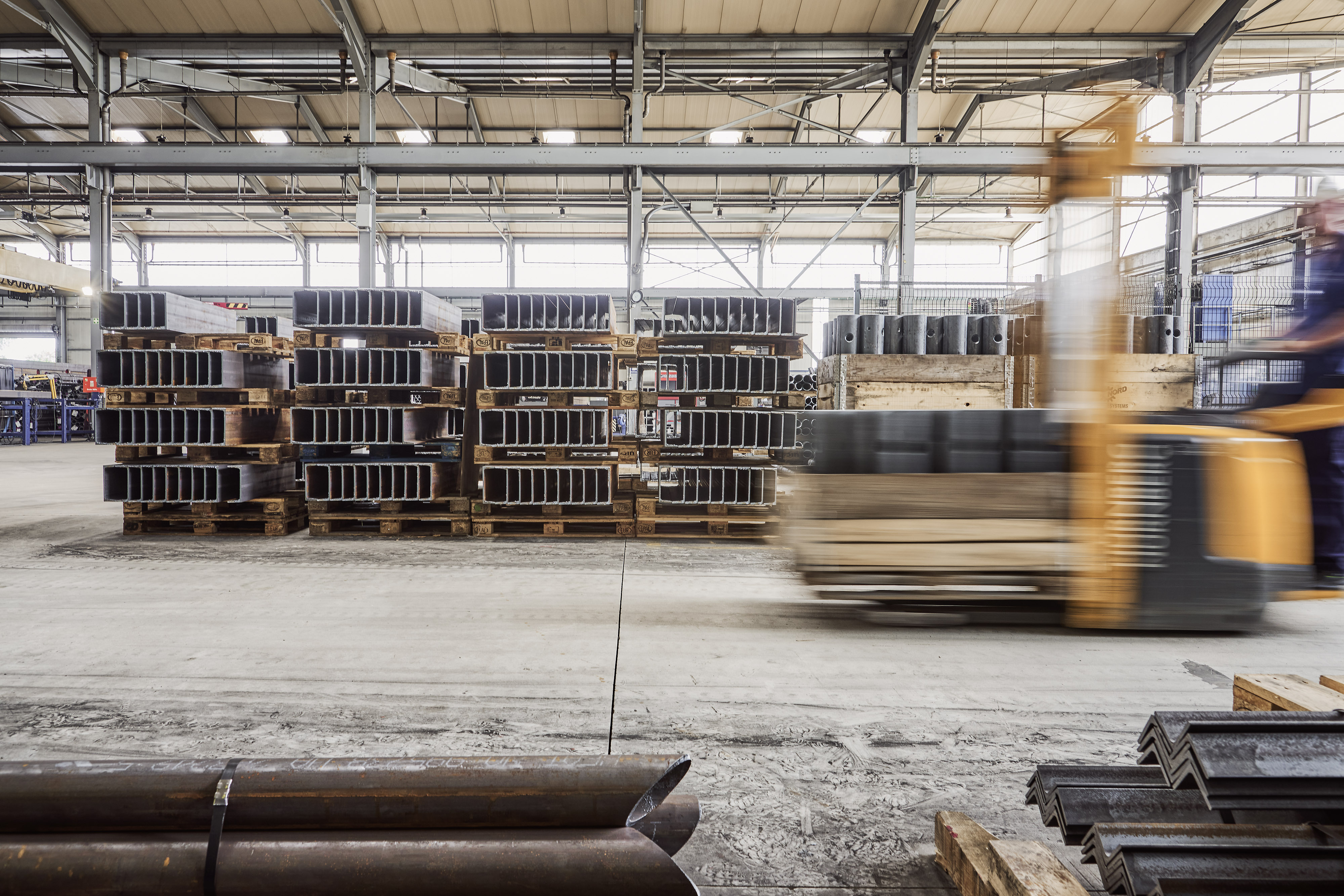 A forklift moves quickly past stacked metal beams in an industrial warehouse, with a worker in motion and steel structures in the background.